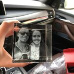 Transparent glass cube engraved with a black-and-white photo of a smiling couple, held in a car interior near the center console and dashboard, with a red gift box nearby.